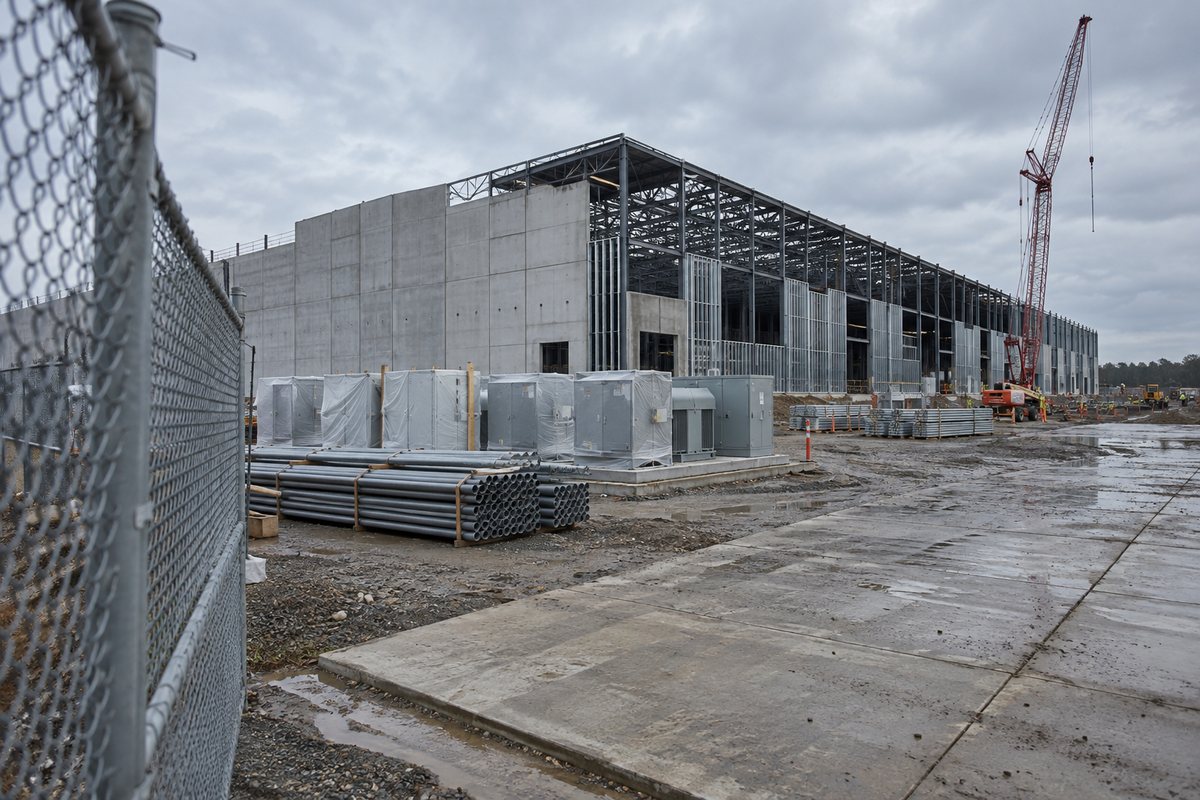 An unfinished industrial building with steel framing, a crane and electrical equipment beside a muddy access road.